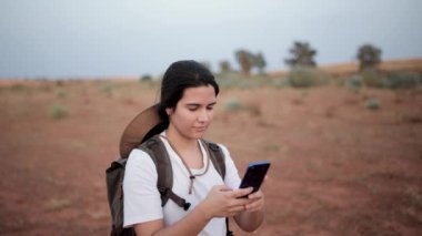 Sırt çantalı memnun bayan gezgin yaz günü çölde dikilirken akıllı telefondan kendini resmediyor.