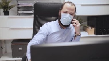 Maskeli genç adam bilgisayarla çalışıyor ve telefonla konuşuyor. 
