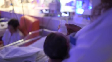 Ekin taşıyan, elinde küçük bir bebek taşıyan ve şişeyle beslenen bir kadının yan görüntüsü. Bağımlılık yapan hastanedeki tıbbi ekipmanlara karşı küçük hastane yataklarının yanında dururken.