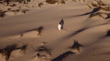 From above drone view of unrecognizable romantic bride and groom in wedding clothes holding hands and strolling on sandy dunes at seaside