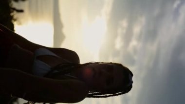 Doğada dans ederken, uzun örgülü saçlarını mavi gökyüzüne doğru sallayan sütyenli enerjik yetişkin bir kadının düşük açılı el kamerası görüntüsü.