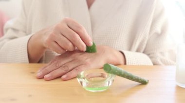 Aydınlık odada günlük cilt bakımı sırasında masada otururken yeşil aloe vera ile elini ovuşturan isimsiz bir kadın.