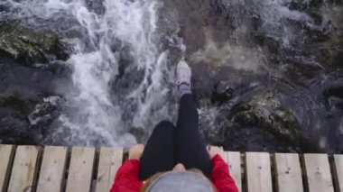Sırt çantalı yorgun turist dağ nehri üzerindeki köprüde sallanan bacaklarıyla oturuyor.