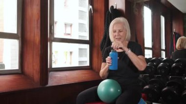 Spor salonundaki halterlere karşı küçük bir egzersiz topuyla bankta otururken şişeden su içen susamış yaşlı bayan sporcu.