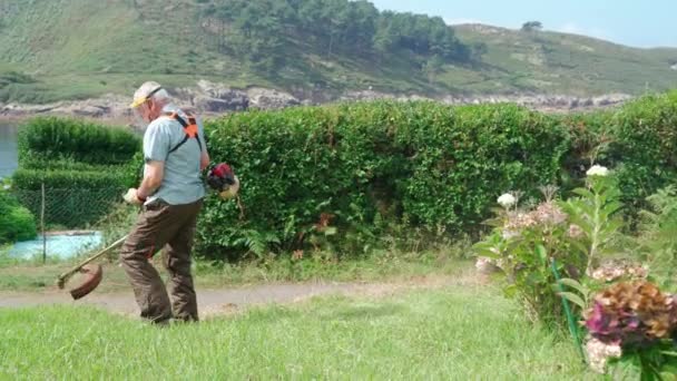 保護シールドマスクの高齢男性 湖畔の芝刈り機で裏庭で草を刈る — ストック動画