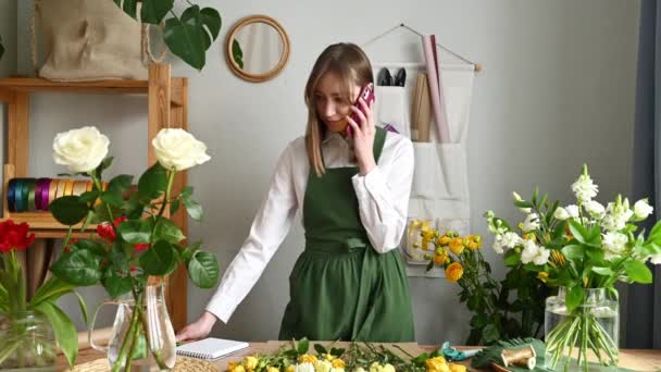 全体的に緑を着て スマートフォンで話しながらメモを作り 様々な花を持つ花屋で注文を受ける陽気な女性 — ストック動画