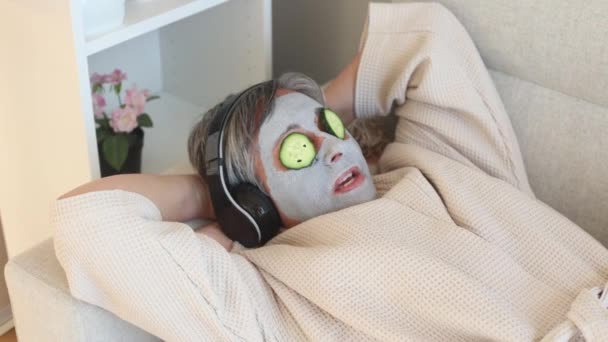 ソファーに横たわりながら音楽を聴く目に反老化粘土マスクとキュウリを持つ上記の女性から — ストック動画