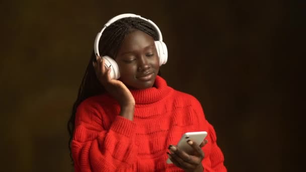 赤色のポジティブな若い黒人女性は スマートフォンを使用して長袖トップをかぎ針編みし ぼやけた茶色の背景でヘッドフォンの音楽を聞いています — ストック動画