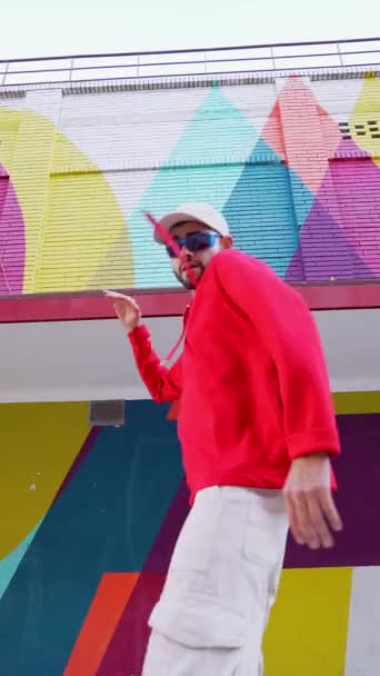 从下面的男性穿着时髦的红色帽衫在城市街道上的五颜六色的建筑物上跳舞 — 图库视频影像