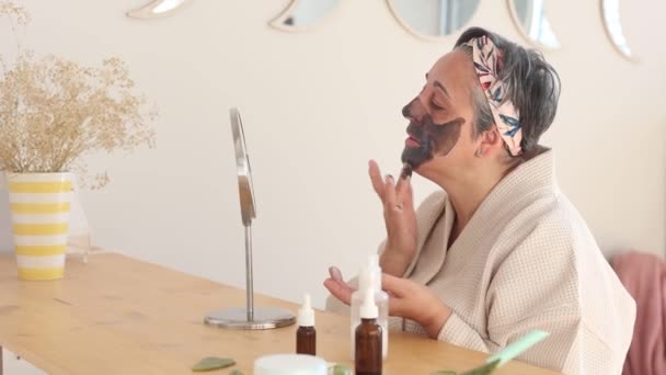 Feminino Roupão Aplicando Máscara Barro Rosto Enquanto Olha Espelho Enquanto — Vídeo de Stock