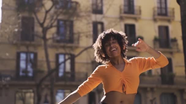 下からハッピーな若いアフリカ系アメリカ人女性のハンドヘルドショット カーリーヘアでオレンジ色のトップとヘッドフォンで音楽を聴きながら 建物に対して街路で踊りながら目を閉じます — ストック動画