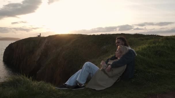 西班牙阿维拉市 穿着外套的男人和女人在日落时坐在悬崖上 彼此靠在一起观察大海的侧视图 — 图库视频影像
