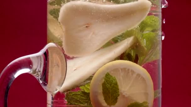 新鮮な熟した梨 レモン ミントのクローズアップショットは 赤い背景に水を投げ込まれるガラス瓶に透明な水で葉を残します — ストック動画