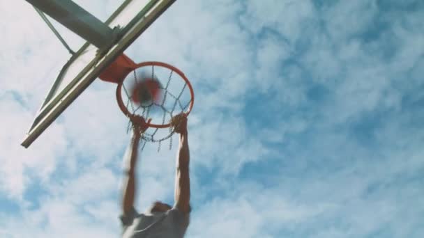 下の民族スポーツマンからスラムダンクシュートを実行し 路上でバスケットボールのトレーニング中に曇った空に向かってフープにぶら下がっている — ストック動画