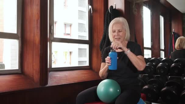体育館のダンベルに対して小さな運動ボールとベンチに座っている間 ボトルから水を飲む喉の渇きシニア女性選手 — ストック動画