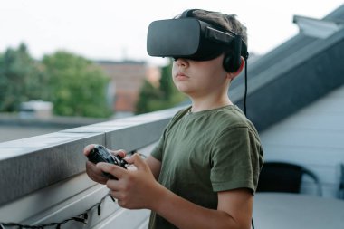 Balkonda tek başına dikilip VR gözlük ve joystick kullanan meraklı küçük çocuk.