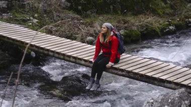 Sırt çantalı yorgun turist dağ nehri üzerindeki köprüde sallanan bacaklarıyla oturuyor.