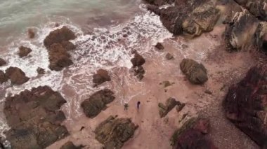 Kayalık kayalıklarla kumsalda duran, güneşli bir günde dalgalı deniz suyuna bakan ve İrlanda 'da vakit geçiren isimsiz insanların hava görüntüsü.
