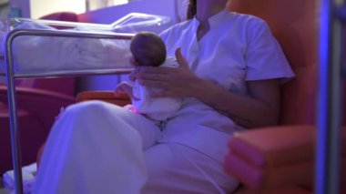 Doktor koltukta oturuyor ve yeni doğan bebeği şişeden besliyor. Hastanedeki ekipmanlarla odada kafalarını tutuyor.