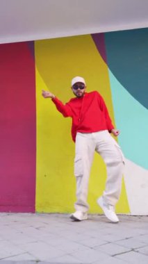 Şehir caddesindeki renkli binaya karşı dans eden şık kırmızı kapüşonlu bir erkekten.