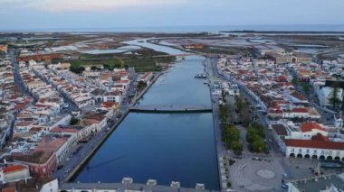 Portekiz 'in Algarve şehrinde bulutlu günbatımına karşı Tavira kasabasında akan yaya köprüleriyle sakin nehir manzarası.