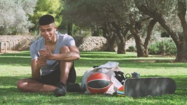 Spor kıyafetli, kablosuz kulaklıklı, akıllı telefondan mesajlar okuyan ve çimlerin yanında oturmuş parkta dinlenirken kıkırdayan genç bir etnik adam.