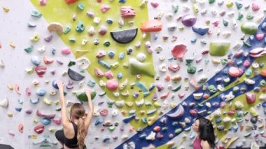 Spor giyim sektöründe kadın dağcı, spor salonunda antrenman yaparken yerde rehberlik ederken parlak tutacaklarla kaya duvarına tırmanıyor.