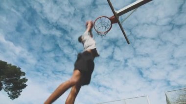 Alttaki etnik sporcudan basket antrenmanında basket potasına sıkıca sarkıyor ve bulutlu gökyüzüne karşı potaya tutunuyor.