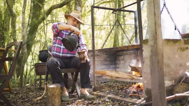 アルゼンチンの男性は 田舎で燃える火の上の棚に対してカラバッシュグルドにケトルから熱い水を注いで — ストック動画