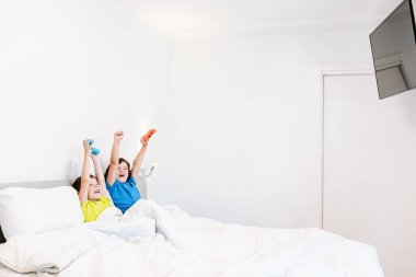 İçerikli kardeşler heyecan içinde kollarını kaldırırken aynı zamanda oyun oynarken yatak odasındaki battaniyenin altında rahat bir yatakta oturuyorlar.