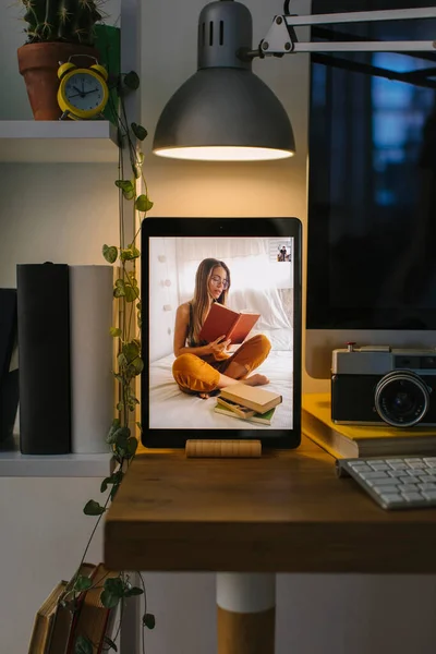 Girl reading in her bed during a video call in Corona Times. The tablet is in a front view over a desk with a computer, some books and a camera.