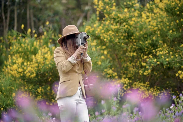 Female photographer in hat with closed eyes filming video with retro video camera while standing near trees with blooming yellow flowers in daylight