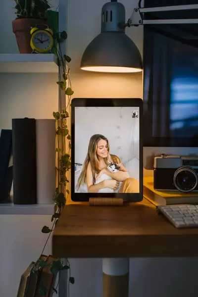 Girl looking at her cat in the bed during a video call with a friend in Corona quarantine. The tablet is in a front view over a desk with a computer, some books and a camera.