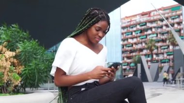 Şık, Afro-Amerikan kadın, gündelik kıyafetlerle ve uzun örgülü saçlarıyla bankta oturarak akıllı telefonlarda çağdaş binalara göz atıyor.