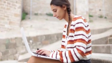 Genç kız öğrenci internette geziniyor ve şehirde sırt çantasının yanında beton basamaklarda otururken ödev yapıyor.