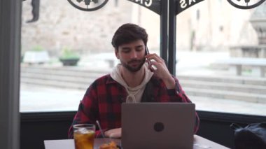 Kendine güvenen sakallı erkek, modern bir netbook ve aydınlık kafede, pencerenin kenarında otururken telefonla konuşuyor. 