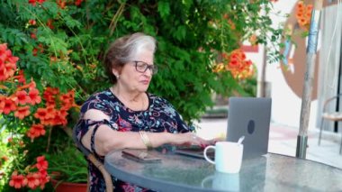 Günlük kıyafetli ve gözlüklü yaşlı bir kadın masada otururken netbook 'un yanında çay tabağı ve bahçede çiçekli yeşil bitkiler üzerinde sörf yapıyor.