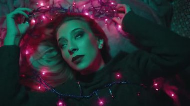 From above of relaxed young female with Christmas reindeer horns and earphones listening to music while lying on bed with glowing garland in dark bedroom with neon illumination