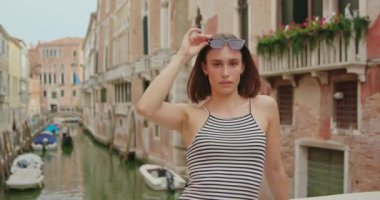 Çizgili bluzlu ve güneş gözlüklü neşeli genç bir kadın Venedik, İtalya 'da hafta sonu gondollarıyla köprü kenarına yaslanıyor.