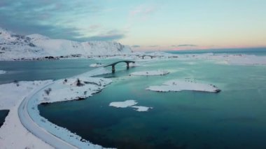 Kışın gün batımında karlı sahildeki kırsal yol ve donmuş deniz suyunun üzerindeki köprü manzarası.