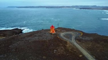 İzlanda 'da fırtınalı bir günde dalgalanan denizin kıyısındaki uçurumun kenarındaki kırmızı deniz feneri binasının drone görüntüsünün etrafında dön