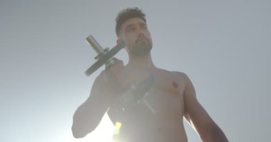 Çıplak gövdeli yakışıklı erkek sporcu yazın mavi gökyüzüne karşı dururken dambıllarıyla egzersiz yapıyor.
