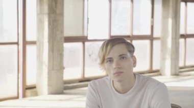 Büyük pencereli ve camdan çatılı boş bir salonda oturan ve kameraya bakan düşünceli genç adam.