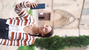 Günlük kıyafetlerle gülümseyen genç bir kadın şehir caddesinde cep telefonundan video görüşmesi yapıyor.