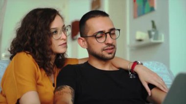 Kanepede otururken ve dizüstü bilgisayar başında video görüşmeleri yaparken gündelik giysiler ve gözlük takan neşeli genç çift.