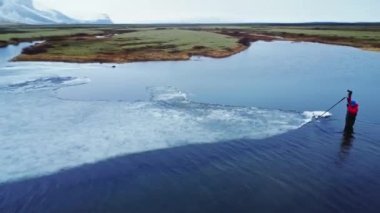 Soğuk nehirde dikilen ve gri günde İzlanda 'da fotoğraf makinesiyle tripodu takan dış giysili gezginin drone görüntüsünü yakınlaştır