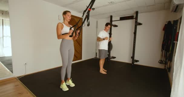 在宽敞的体育馆与教练进行个人训练的过程中 身材瘦弱的女运动员在Trx设备上做隆起动作 — 图库视频影像
