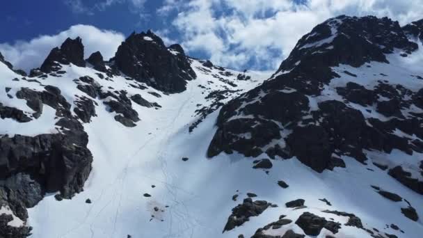 阳光明媚的日子里 远处滑雪者在雪地的斜坡上滑行 四周都是黑色的岩石 — 图库视频影像