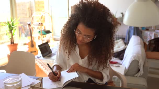 現代の若いアフリカ系アメリカ人女性リモート従業員カジュアルな衣装でテーブルに座ってノートパソコンと家のオフィスで働いている間プランナーで書く — ストック動画