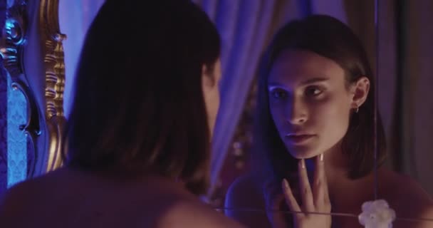 若い女性は 自宅で夜の不明瞭な部屋で鏡を見ながらイヤリングとダークヘアを調整する — ストック動画